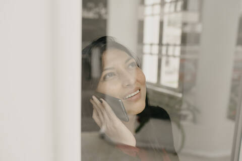 Portrait of smiling woman using smartphone behind window pane stock photo