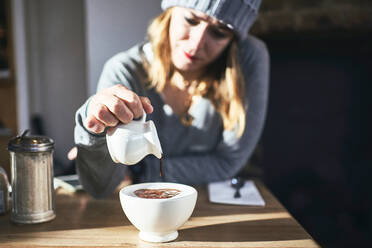 Junge Frau in warmem Pullover und Hut gießt Schokoladensirup in einen weißen Becher mit heißem Getränk, während sie an einem Tisch in einem sonnigen Café sitzt - ADSF09106