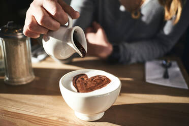 Abgeschnittene, nicht erkennbare Frau in warmem Pullover und Hut gießt Schokoladensirup in einen weißen Becher mit heißem Getränk, während sie an einem Tisch in einem sonnigen Café sitzt - ADSF09107