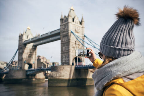 Rückenansicht einer weiblichen Reisenden in warmer Kleidung und Hut, die die Tower Bridge mit ihrem Smartphone während einer Stadtbesichtigung in London an einem sonnigen Tag fotografiert - ADSF09108