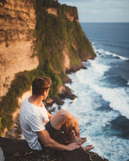 Seitenansicht eines Mannes, der allein auf einer Klippe in der Höhe sitzt und auf eine majestätische Landschaft mit Meereswellen blickt, Bali - ADSF09109