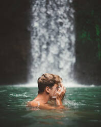 Side view of man washing face while swimming in clear water of lake with waterfall on background, Bali - ADSF09112