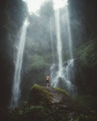 Blick von unten auf einen Mann, der auf einem Felsen zwischen hohen grünen Klippen mit Wasserfall steht, Bali - ADSF09119