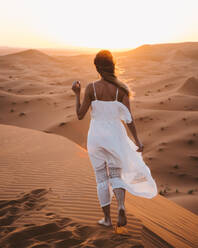 Rückenansicht einer barfuß gehenden Frau im weißen Sommerkleid auf einer Sanddüne in der endlosen Wüste bei Sonnenuntergang, Marokko - ADSF09128