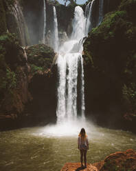 Back view of woman standing in desert terrain with powerful majestic waterfall on green cliffs, Morocco - ADSF09132