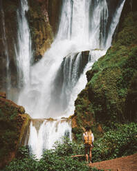 Back view of woman standing in desert terrain with powerful majestic waterfall on green cliffs, Morocco - ADSF09133