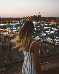 Rückenansicht einer trendigen Frau mit fliegenden Haaren, die auf einem Balkon vor den hellen Lichtern einer marokkanischen Stadt in der Dämmerung steht - ADSF09134