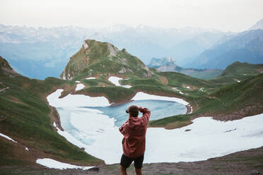 Back view of anonymous male tourist taking photo of mountain lake surrounded by snow and ridge in Pyrenees - ADSF09137