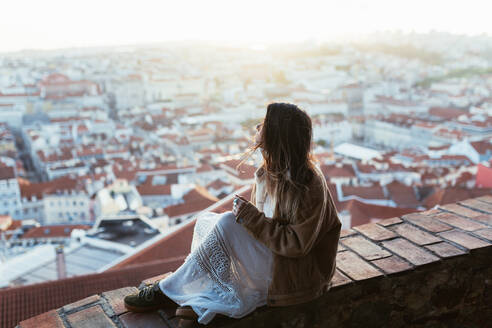 Seitenansicht einer jungen Frau in Jacke und Kleid, die auf einer Mauer sitzt und das Stadtbild an einem sonnigen Morgen in Portugal bewundert - ADSF09149