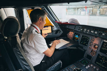 Seitenansicht eines männlichen Flugkapitäns, der im Cockpit eines modernen Flugzeugs sitzt und die Flugdokumente liest, während er sich auf den Abflug vorbereitet - ADSF09156