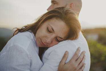 Seitenansicht des fürsorglichen liebenden Paares in passenden weißen Hemden, die sich vertrauensvoll umarmen, während sie auf einem grasbewachsenen Feld bei Sonnenuntergang stehen - ADSF09181