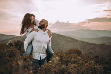 Loving couple in matching white shirt having fun and riding piggyback at grassy mountain slope - ADSF09182