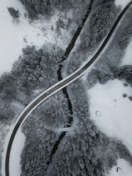 Aerial view of asphalt road stretching over river Ach flowing through snow-covered forest - MALF00054