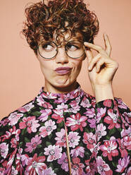 Thoughtful doubtful beautiful curly haired female in trendy eyeglasses and stylish colorful blouse with floral ornament looking away against pink background - ADSF09205