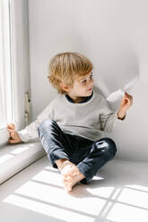 Happy blonde little child in casual clothes playing with paper airplane while sitting barefoot on window sill on sunny day - ADSF09216