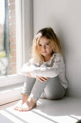 Verärgertes kleines Mädchen mit lockigen Haaren, das einen grauen, gemütlichen Schlafanzug trägt und unzufrieden in die Kamera schaut, während es mit verschränkten Armen auf der Fensterbank sitzt - ADSF09217