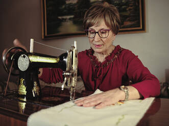 Ältere Dame mit Brille benutzt eine Retro-Nähmaschine, um eine Leinenserviette in einem gemütlichen Zimmer zu Hause zu nähen - ADSF09223