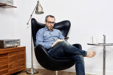 Mann liest ein Buch und trinkt entspannt einen Kaffee im Sessel zu Hause - ADSF09225