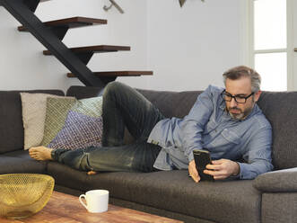 Man using phone alone on sofa in living room at home - ADSF09232