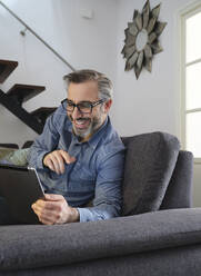 Smiling Man using tablet surfing the internet alone on the couch in the living room at home - ADSF09233
