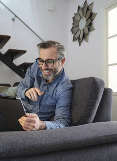 Lächelnder Mann, der mit einem Tablet im Internet surft, allein auf der Couch im heimischen Wohnzimmer - ADSF09233