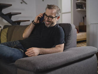 Entspannter Mann telefoniert lächelnd allein auf dem Sofa im Wohnzimmer zu Hause - ADSF09237