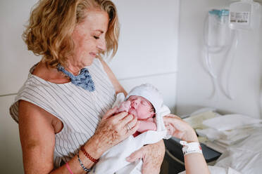 Glückliche reife Frau, die ihr neugeborenes Enkelkind auf der Krankenstation begrüßt, während die junge Mutter im Bett liegt und medizinisch behandelt wird - ADSF09250