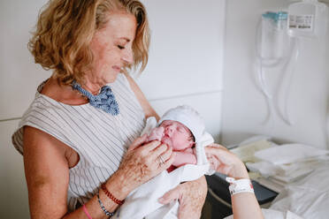 Happy mature female greeting newborn grandchild in hospital ward while young mother lying in bed and receiving medical treatment - ADSF09250