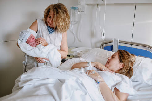 Glückliche reife Frau, die ihr neugeborenes Enkelkind auf der Krankenstation begrüßt, während die junge Mutter im Bett liegt und medizinisch behandelt wird - ADSF09252
