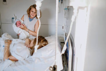 Glückliche reife Frau, die ihr neugeborenes Enkelkind auf der Krankenstation begrüßt, während die junge Mutter im Bett liegt und medizinisch behandelt wird - ADSF09253