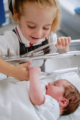 From above of cheerful curious little girl standing near newborn baby sibling sleeping in hospital cradle in ward - ADSF09256