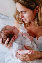 From above closeup of loving mother holding adorable newborn baby while sitting on bed at home - ADSF09259