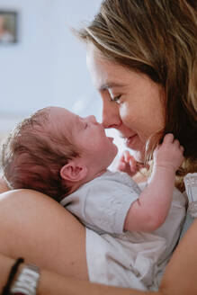 Nahaufnahme einer liebenden Mutter, die ein bezauberndes neugeborenes Baby hält, während sie auf dem Bett zu Hause sitzt - ADSF09264