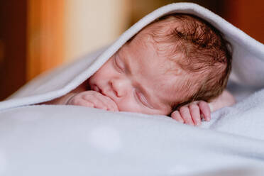 Calm cute infant with closed eyes covered with blanket resting in bed against blurred interior of light bedroom - ADSF09267