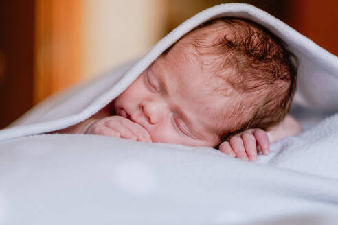 Ruhiges niedliches Kleinkind mit geschlossenen Augen, das mit einer Decke zugedeckt im Bett liegt, vor einem unscharfen Interieur eines hellen Schlafzimmers - ADSF09267