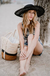 Hübsche blonde Frau mit schwarzem Hut sitzt mit Sommertasche im Sand und schaut in die Kamera - ADSF09269