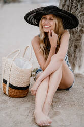 Hübsche blonde Frau mit schwarzem Hut sitzt mit Sommertasche im Sand und schaut weg - ADSF09270
