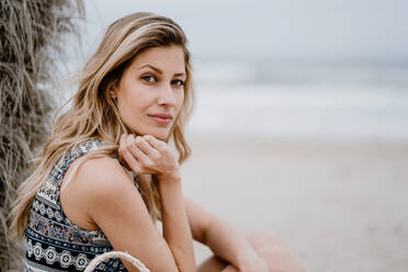 Porträt einer jungen, schönen, blonden, verführerischen Frau, die am Strand sitzt und in die Kamera schaut - ADSF09277
