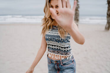 Fröhliche blonde Frau in buntem Oberteil und Jeansshorts, die ihr Gesicht mit der Hand am Meeresstrand abdeckt - ADSF09280