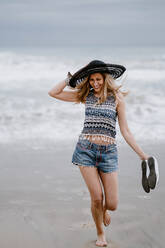 Attraktive Frau mit schwarzem Hut, die eine Strandtasche und Schuhe hält, während sie den malerischen Blick auf das Meer genießt - ADSF09281