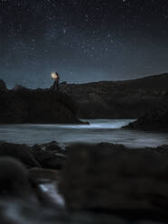 Silhouette of traveler standing with lantern on rocky shore of river and admiring mysterious scenery of tranquil water during starry night in highlands - ADSF09286