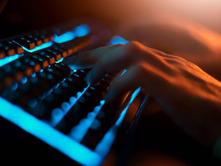 Hoher Winkel des unerkennbaren Mannes in Freizeitkleidung, der auf einer schwarzen Tastatur mit bequemer blauer Hintergrundbeleuchtung tippt, während er einen Computer in einem dunklen Raum mit gedämpftem Licht bei Nacht benutzt - ADSF09288