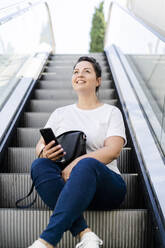 Lächelnde kurvige junge Frau sitzt auf Rolltreppe mit Handy und schaut nach oben - GIOF08629