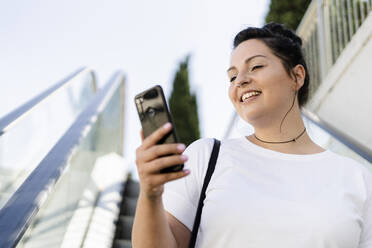 Lächelnde kurvige junge Frau, die auf einer Rolltreppe ein Mobiltelefon benutzt - GIOF08630