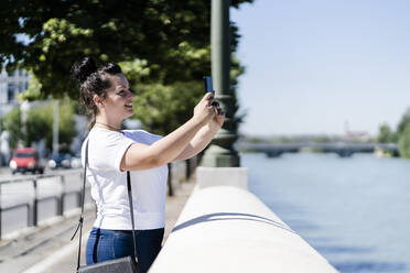 Lächelnde junge Touristin, die ein Selfie am Flussufer macht - GIOF08633