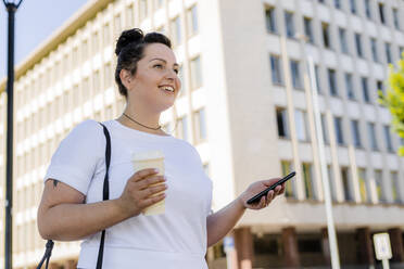 Smiling curvy young woman holding mobile phone in the city - GIOF08644