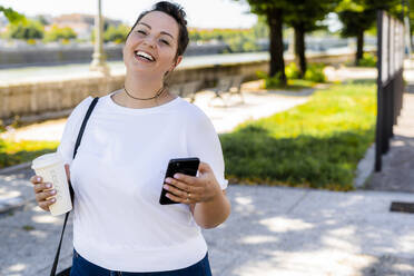 Portrait of a happy curvy young woman with takeaway drink and mobile phone in the city - GIOF08646