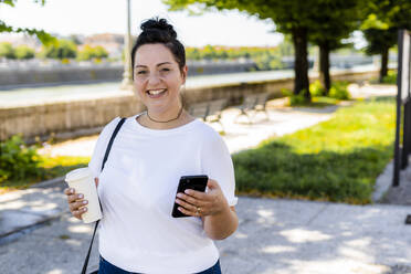 Porträt einer lächelnden, kurvenreichen jungen Frau mit einem Getränk zum Mitnehmen und einem Mobiltelefon in der Stadt - GIOF08647