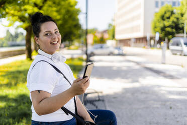 Portrait of a smiling curvy young woman with mobile phone sitting on a bench in the city - GIOF08648