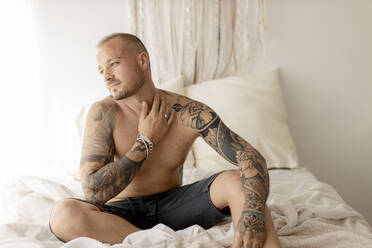 Shirtless hipsterwith tattoo on arms sitting on bed - GMLF00408